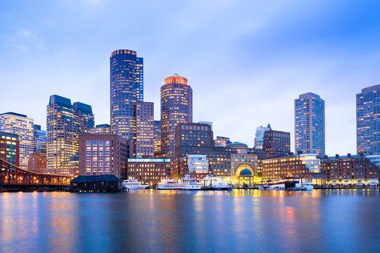 Photograph of the Boston skyline at dusk seen across the harbour, the lit towers reflected in the water