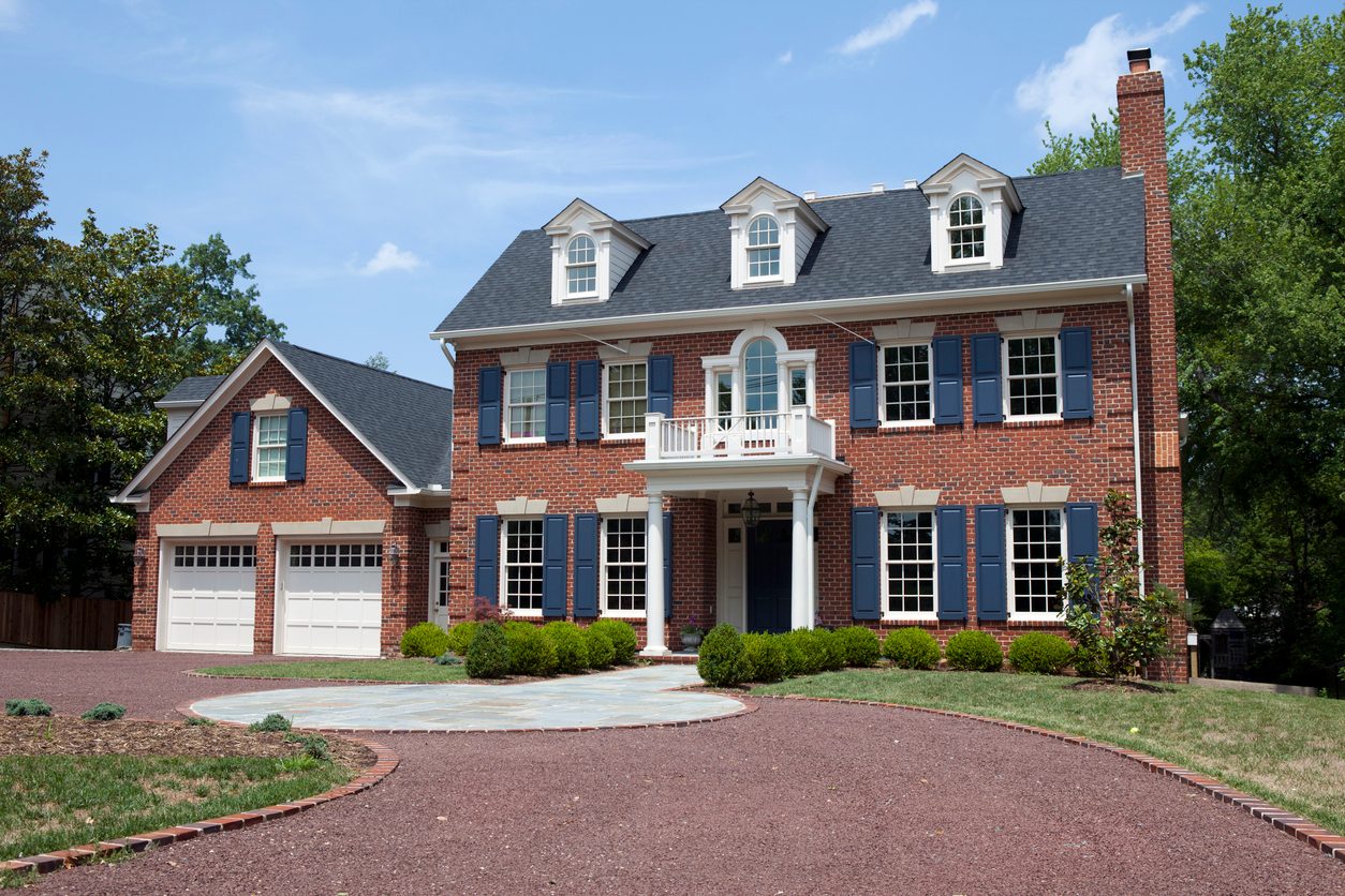 Photograph of a two-storey red brick Colonial house with blue shutters, three gabled dormers, an entry portico with a balcony over it, and a two-car garage wing