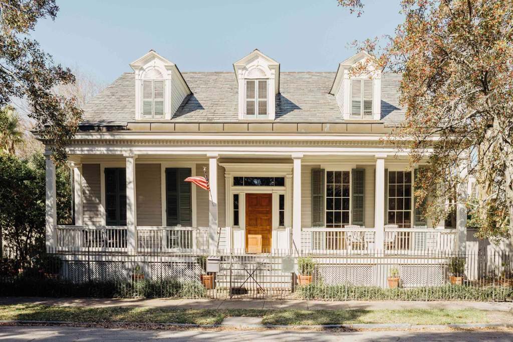Photograph of a broad one-storey cream house with a low hipped roof, three pedimented dormers and a full-width raised veranda on square columns