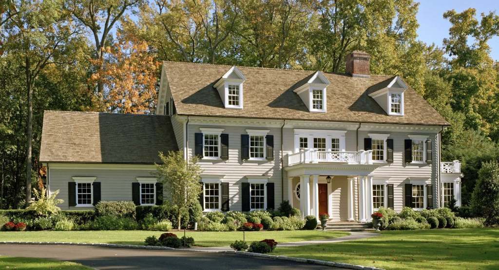 Photograph of a cream clapboard Colonial house with black shutters, three gabled dormers, an entry portico with a balcony over it, and a lower one-storey wing on the left